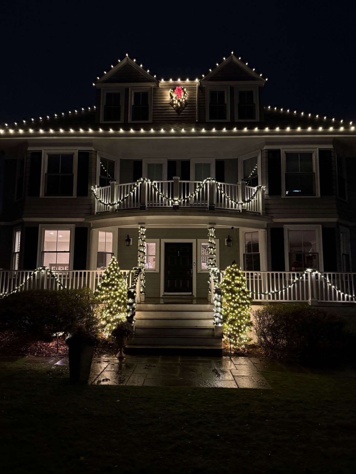 Colonial house lit up by ReFresh Holiday Lighting: warm white roofline lights, garland on the porch and balcony railings, wrapped columns, and two lit trees by the steps